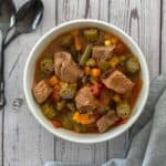 An overhead shot of a white bowl of beef stew with frozen vegetables with black spoons on the side on a wooden table.