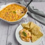 A plate with a serving of corn dip with cream cheese and rotel and chips and the pan of the corn dip in the background with a black spoon in it.