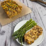 A serving of meatloaf with elbow macaroni with roasted asparagus on a white plate with the rest of the meatloaf in the background on a piece of parchment paper.
