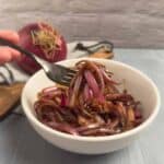 A fork held up with balsamic onion jam on it with a white bowl of the onion jam in the background.
