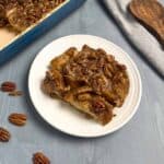 A piece of French toast casserole with brown sugar on a white plate with the casserole dish on the side.