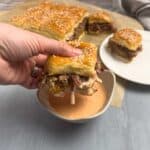 A hand holding a smash burger and dipping it in a small bowl of sauce with more smash burgers in the background.